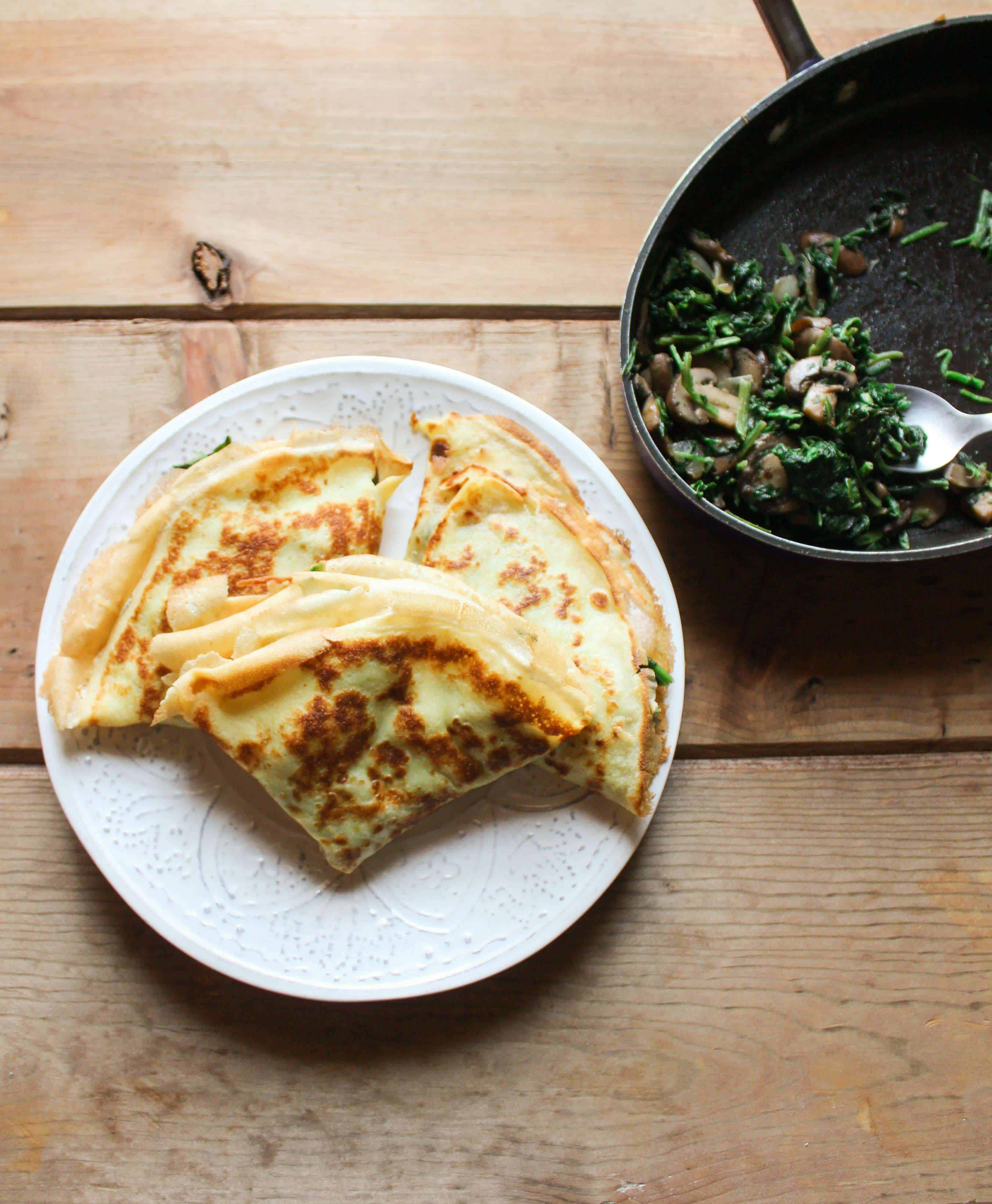 Crepe on a place with a pan of spinach, mushroom, and onion next to it. 