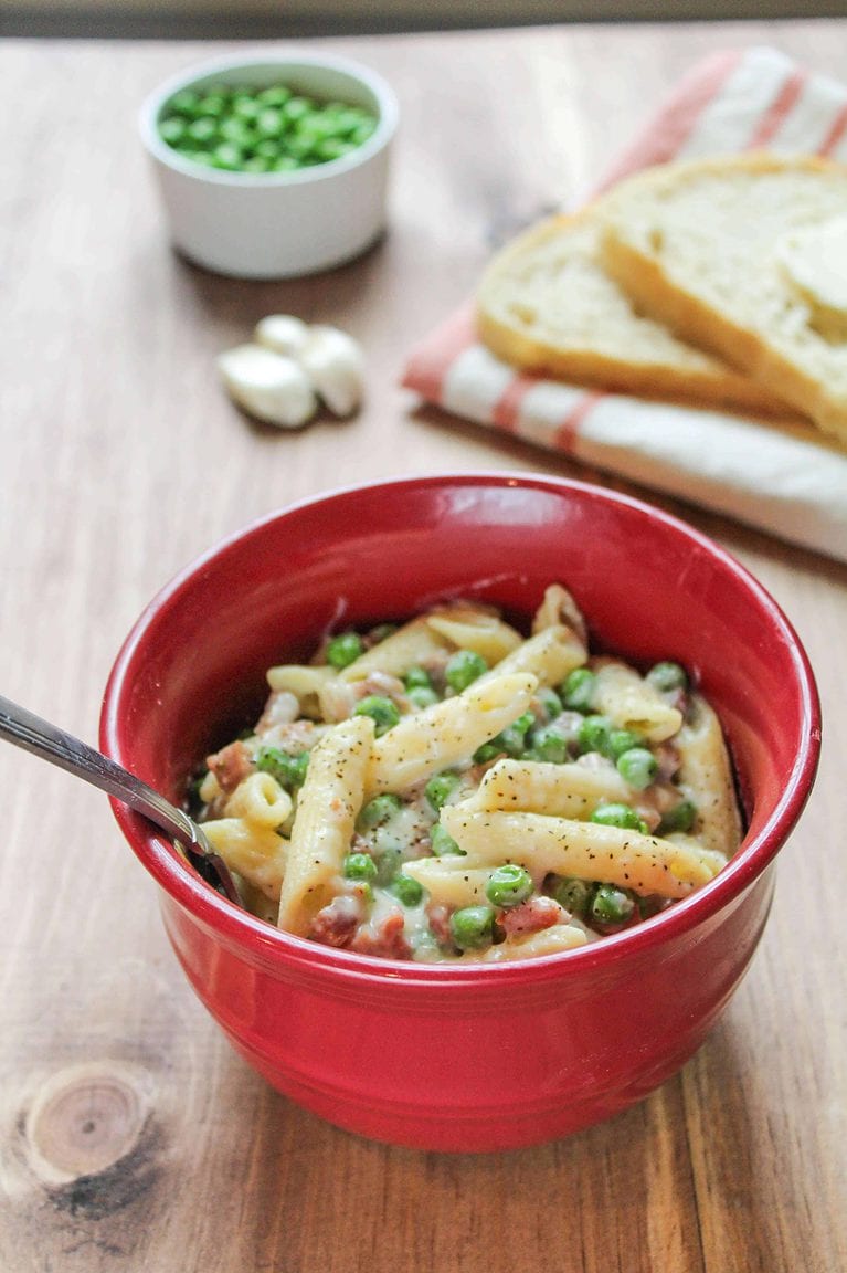 Prosciutto & Pea Alfredo with Penne in a red bowl. Bread and peas are behind it.
