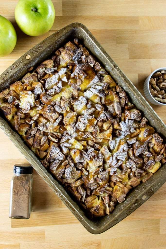 Baked Apple French Toast surrounded by apples, pecans, and a jar of cinnamon 