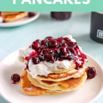 Pancakes topped with whipped cream and Berry Sauce. Next to a jar of berry sauce and a coffee mug