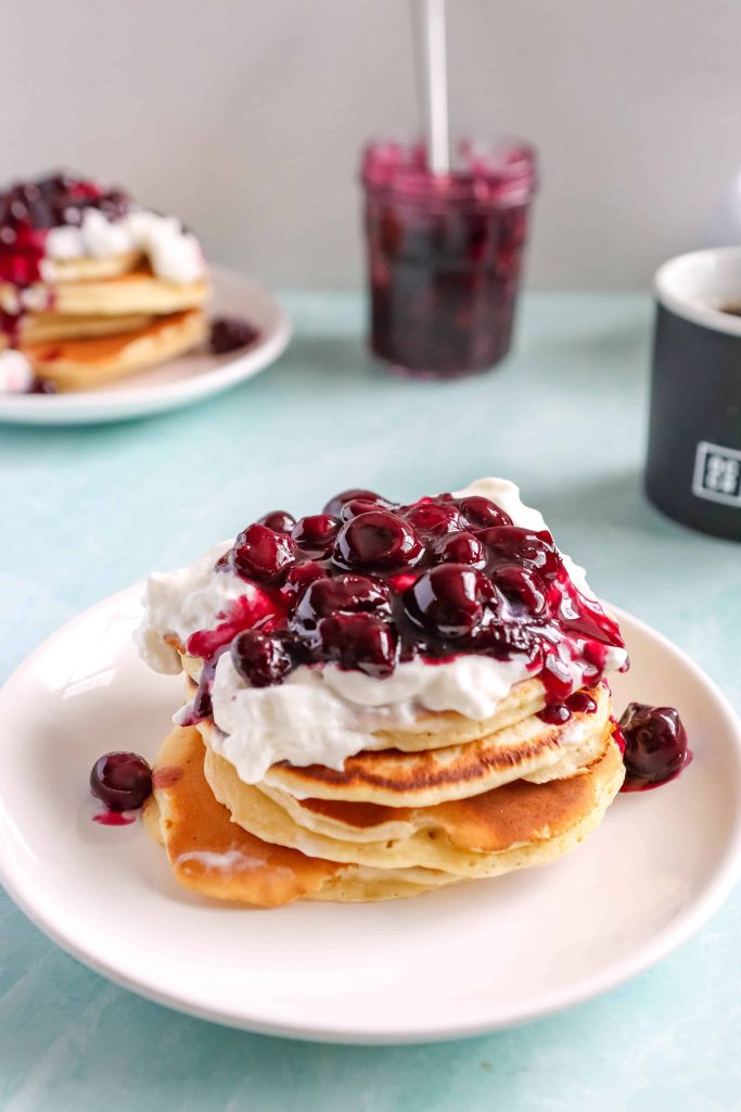 Pancakes topped with whipped cream and  Berry Sauce. Next to a jar of berry sauce and a coffee mug
