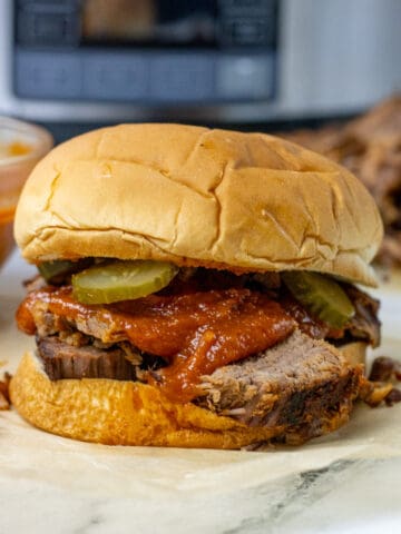 bbq brisket sandwich with bbq sauce and pickles on it. there is a slow cooker in the background
